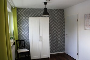 Habitación con armario blanco, una silla, lámpara colgante y puerta con papel tapiz estampado.