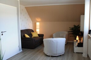 Un espacio habitable pequeño con un sofá oscuro, un sillón blanco y una mesita con lámpara.