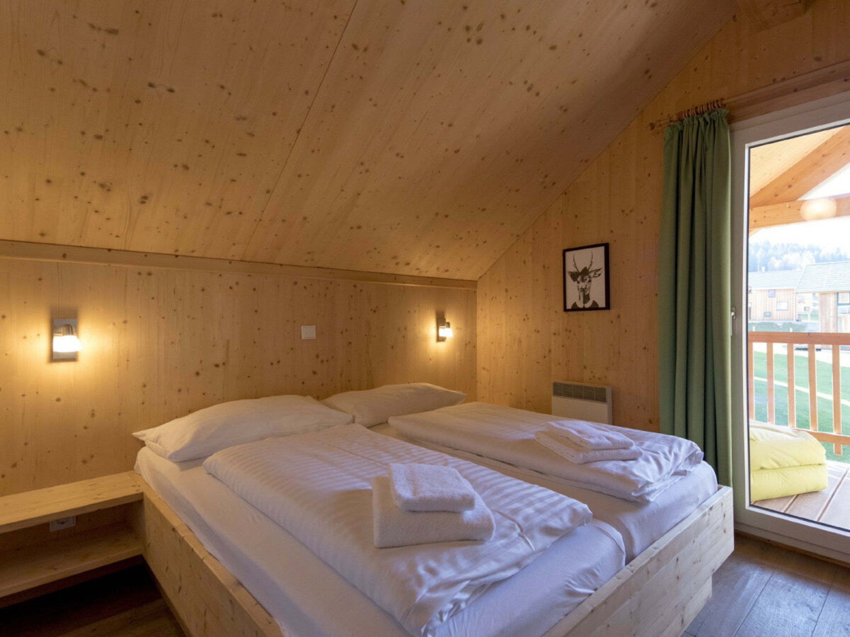 Une pièce en bois avec un lit, deux lampes fixées au mur, un tableau encadré et une fenêtre avec des rideaux.