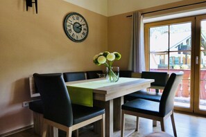 A dining area with a wooden table, black chairs, a clock on the wall, and a window with a view of buildings. - Unterbrandnerlehen FeWo Königssee (Königssee)