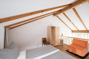 Een slaapkamer met een schuine kap, een bed, een oranje stoel, een houten garderobe en een raam met uitzicht.