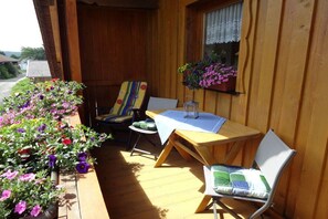 A wooden deck with a table and two chairs, a window with lace curtains, and potted plants with flowers. - Holiday apartment in Böbrach with Wi-Fi (Böbrach)