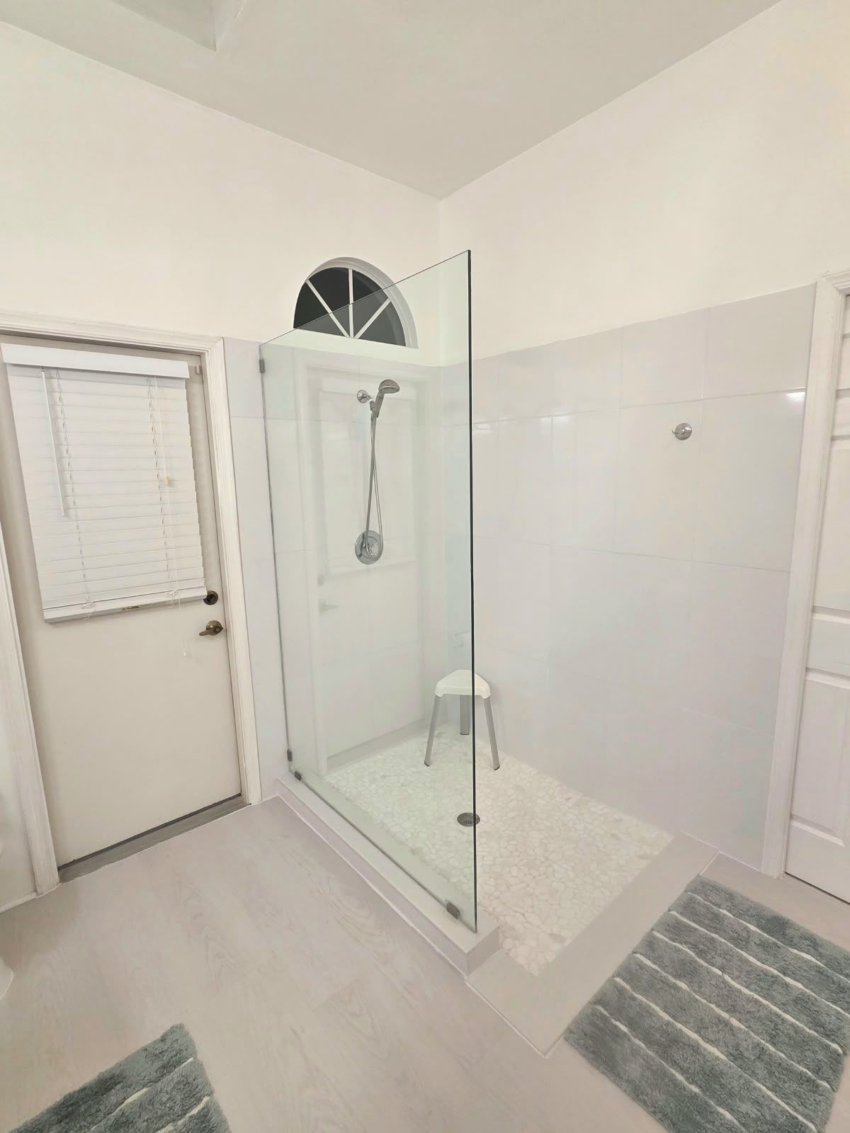 A glass-enclosed shower with a white tiled wall, a small stool, and a white door with a window.