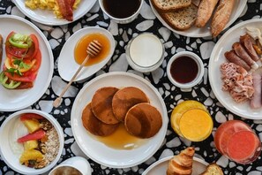 Uma mesa de café da manhã com panquecas, torradas, ovos, bacon e diversas frutas e bebidas.