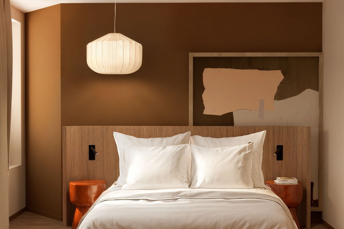 A modern bedroom with a large bed, wooden headboard, a hanging light fixture, and a painting on the wall.