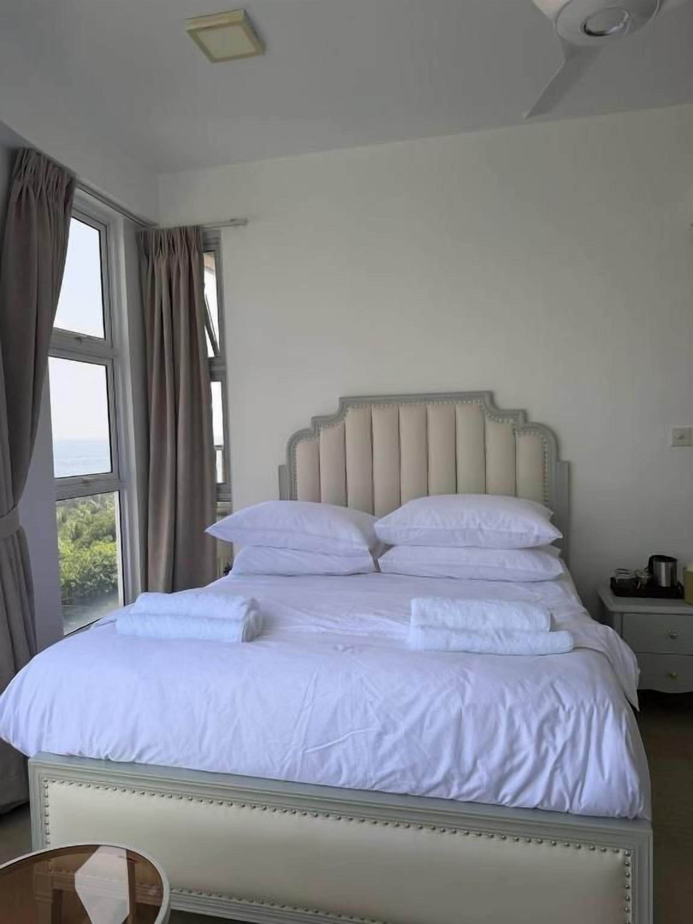 Cama bem arrumada com lençóis e almofadas brancas, mesa de cabeceira com abajur e janela com cortinas.