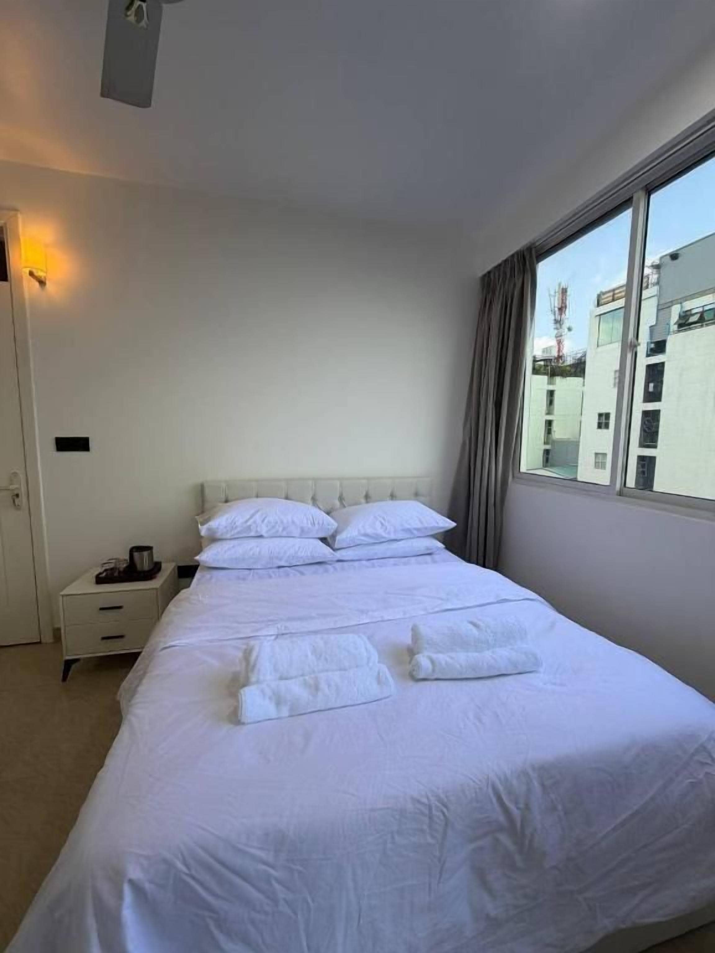 A hotel room with a large bed, a nightstand, a window with curtains, and a view of buildings outside.