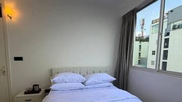 A hotel room with a large bed, a nightstand, a window with curtains, and a view of buildings outside.