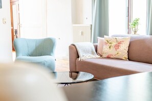 Olohuoneessa on vaaleansininen nojatuoli, ruskea sohva, jossa on kukallisia koristetyynyjä, sekä lasinen sohvapöytä.