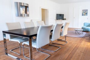 A modern dining area with a wooden table and white chairs, a built-in stove, and a television in the background. - Manor House Burg Vlatten - To the Court (Heimbach)