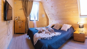 A cozy room with a bed, wooden walls, a window with curtains, a bedside table with a lamp, and a TV mounted on the wall.