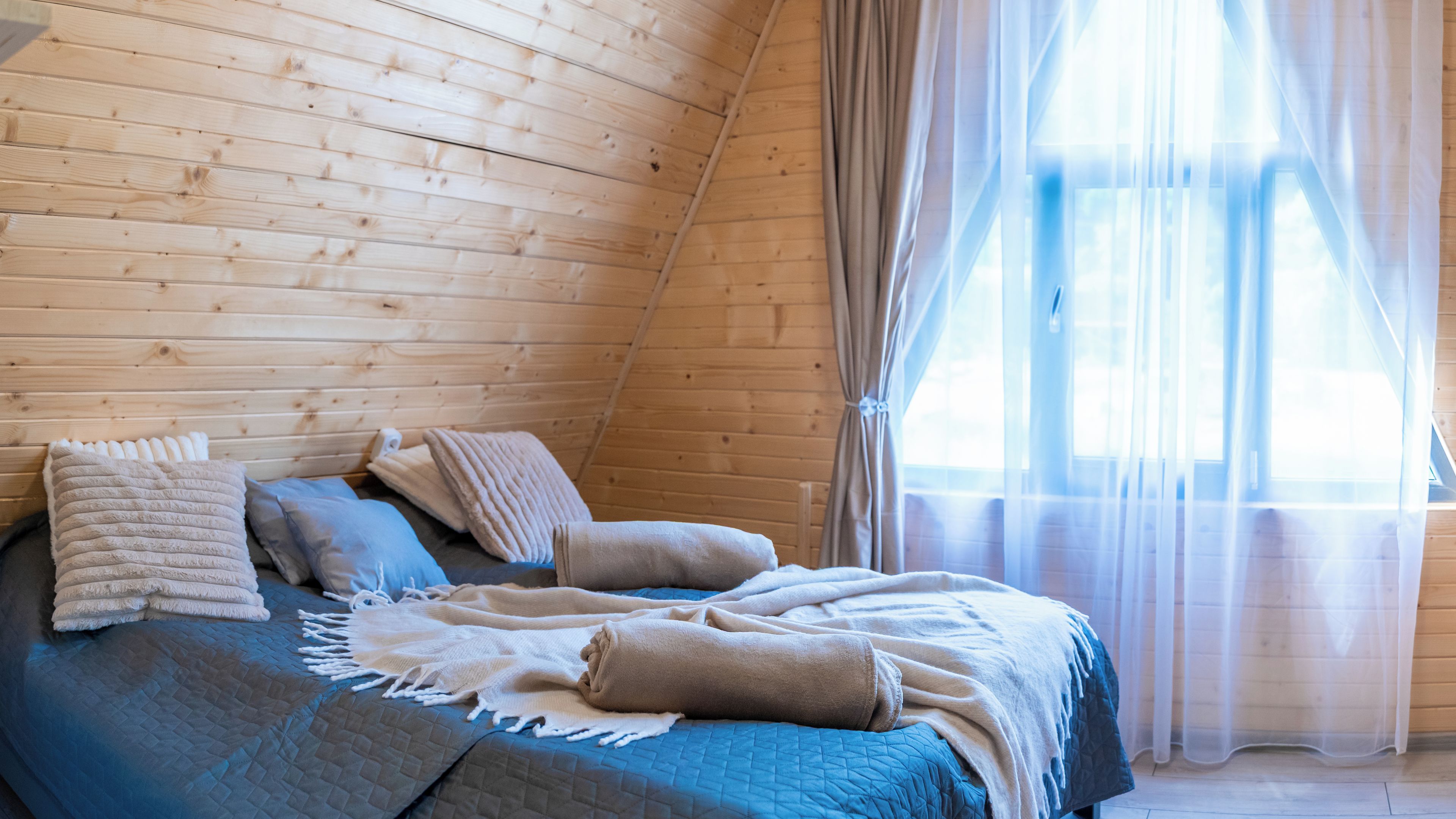 A cozy room with a sloped ceiling, a bed with blue bedding, and sheer curtains on a window.