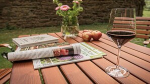Uma mesa de madeira com uma taça de vinho tinto, uma revista, um vaso com flores e maçãs.