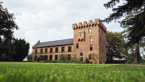 Um grande edifício de tijolos com uma torre imponente, cercado por um gramado bem cuidado e árvores.