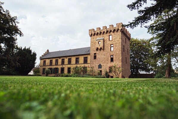 A large brick building with a prominent tower, surrounded by a well-maintained lawn and trees. - Vlatten Manor House - Under the Roof (Heimbach)