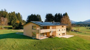 Une maison en bois à deux étages, dotée d’un balcon, située dans un champ verdoyant, avec des arbres et des montagnes en arrière-plan.