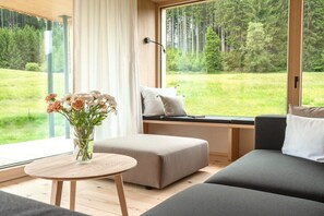 Un intérieur moderne avec un canapé, un pouf et une table agrémentée d’un vase de fleurs. Une fenêtre offre une vue sur un paysage verdoyant.