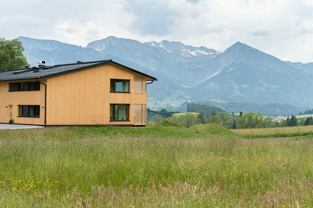 Une maison en bois à deux étages, dotée d’un balcon, qui se dresse sur un fond de montagnes et de champs.
