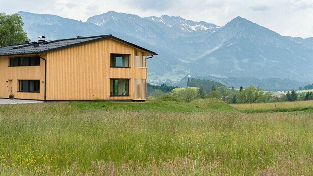 Une maison en bois à deux étages, dotée d’un balcon, qui se dresse sur un fond de montagnes et de champs.