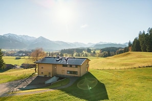 Un bâtiment à deux étages, doté d’un toit en pente, entouré de champs verdoyants et avec des montagnes en arrière-plan.