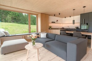 A modern interior with a grey sectional sofa, a round wooden coffee table, and a view of a green landscape through large windows. - Holiday apartment Wald (Bolsterlang)
