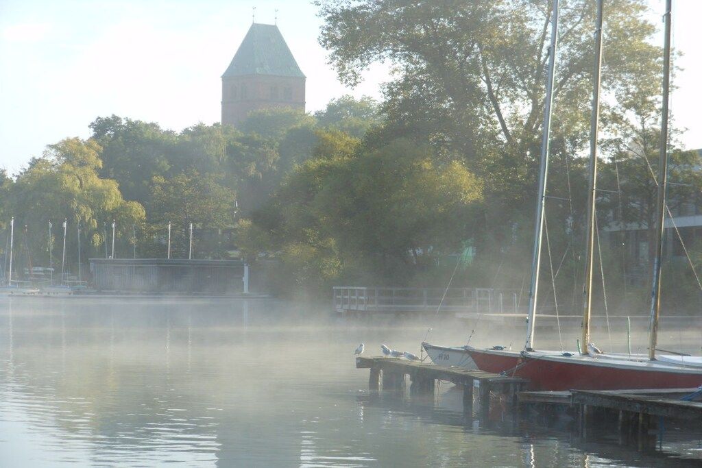 A calm river with boats docked on a pier, surrounded by trees and a distant building.