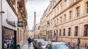 Vista da rua com carros estacionados, uma pessoa caminhando e edifícios com placas.
