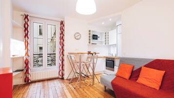 A compact living space with a kitchenette, a dining area with chairs, a sofa with orange cushions, and a red and white patterned curtain.
