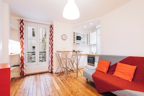 Un espace de vie compact comprenant un coin cuisine, un coin repas avec des chaises, un canapé avec des coussins orange et un rideau à motifs rouges et blancs.