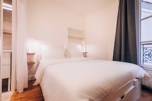 Une chambre avec un grand lit, du linge de lit blanc, un plancher en bois, une table de chevet avec une lampe et une fenêtre avec un rideau.