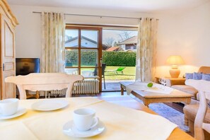 Une pièce comprenant une table à manger dressée pour deux personnes, un canapé, une petite table basse et une vue sur un jardin, visible à travers une porte vitrée.