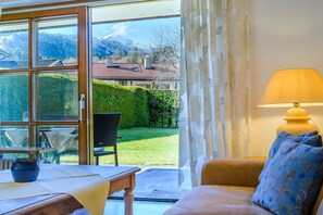 A room with a sofa, a table, and a lamp. There is a view of a garden and mountains outside. - Holiday apartment with 1 bedroom (Garmisch-Partenkirchen)