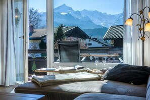 A room with a large window offering a mountain view, a sofa, a table with a glass, and a lamp. - Holiday apartment with 1 bedroom (Garmisch-Partenkirchen)