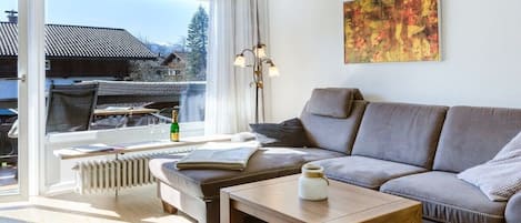 A modern living room with a sofa, a coffee table, and a radiator. There is a window with a view of houses and trees.