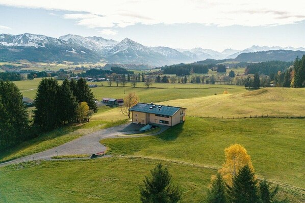 Une maison de campagne avec une allée pavée et un petit espace de stationnement, entourée de champs verdoyants et avec des montagnes en arrière-plan.