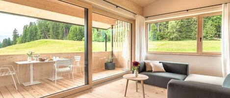 A modern interior with a wooden floor, a sofa, a dining table with chairs, and large windows offering a view of a green landscape.