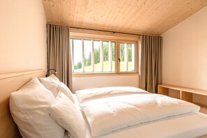 Une chambre avec un plafond en bois, une grande fenêtre avec des rideaux, un lit avec une literie blanche et un banc en bois.