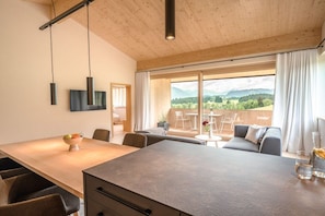 Un intérieur moderne avec un plafond en bois, un coin repas comprenant une table et des chaises, un canapé et une vue sur un paysage montagneux grâce à de grandes fenêtres.