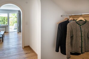 Ein Zimmer mit Holzboden, einem Kleiderständer mit zwei Kleiderbügeln und Blick durch eine gewölbte Tür auf einen Außenbereich mit Tisch und Stühlen.
