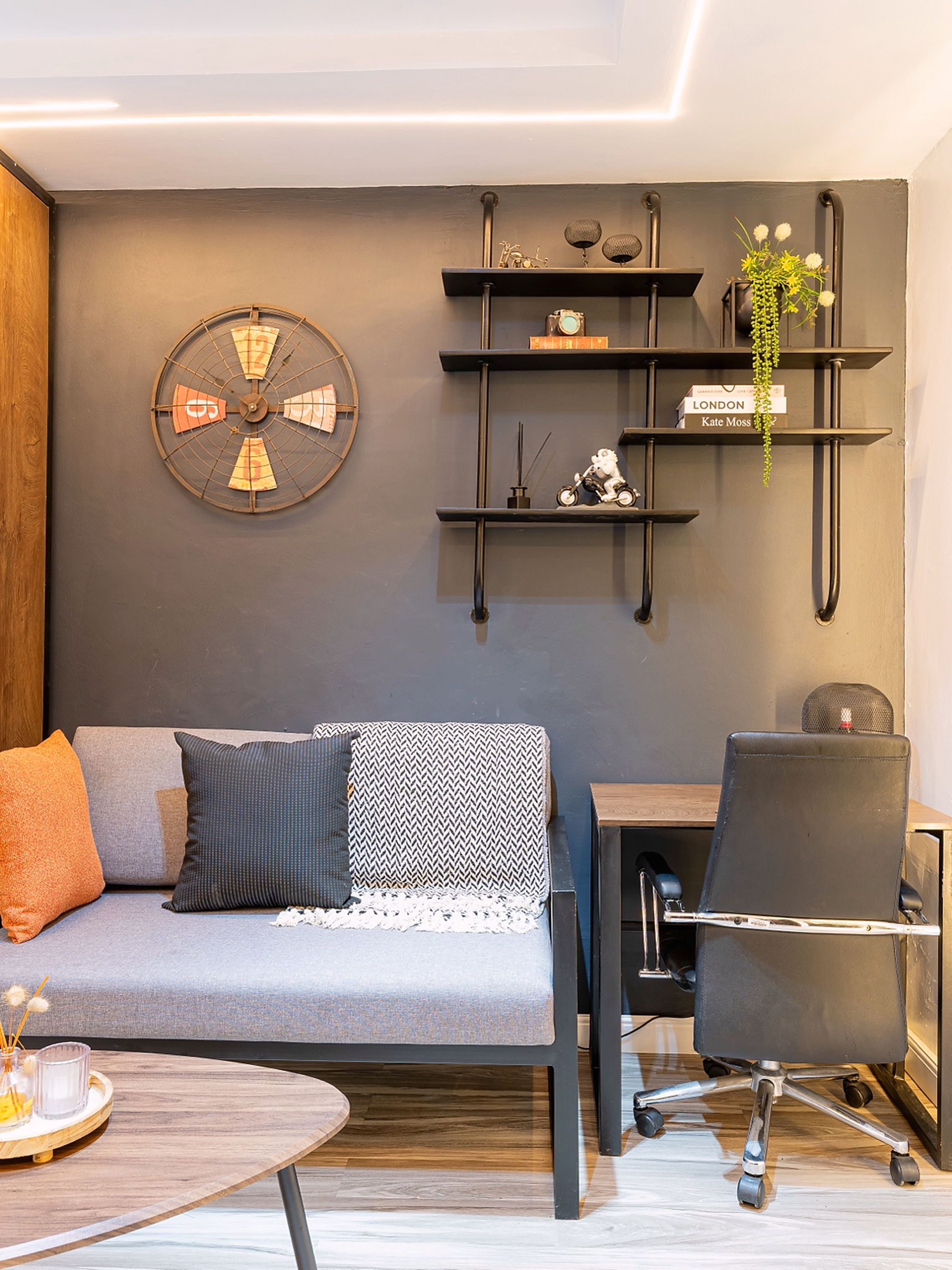 Ein stilvolles Wohnzimmer mit einem grauen Sofa, einer runden Wanduhr, einem kleinen Schreibtisch mit Stuhl und einem Holztisch.