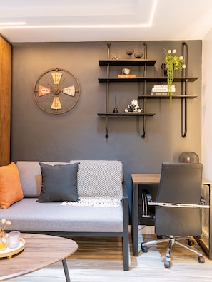 Ein stilvolles Wohnzimmer mit einem grauen Sofa, einer runden Wanduhr, einem kleinen Schreibtisch mit Stuhl und einem Holztisch.