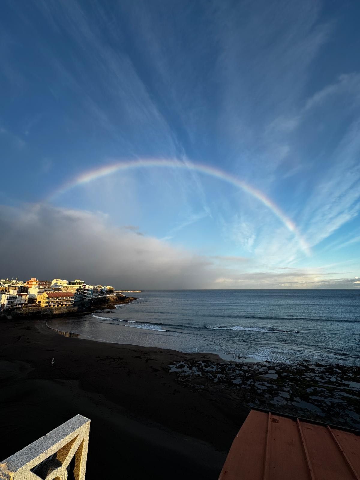 Een uitzicht op de kust met een regenboog, gebouwen en een strand.