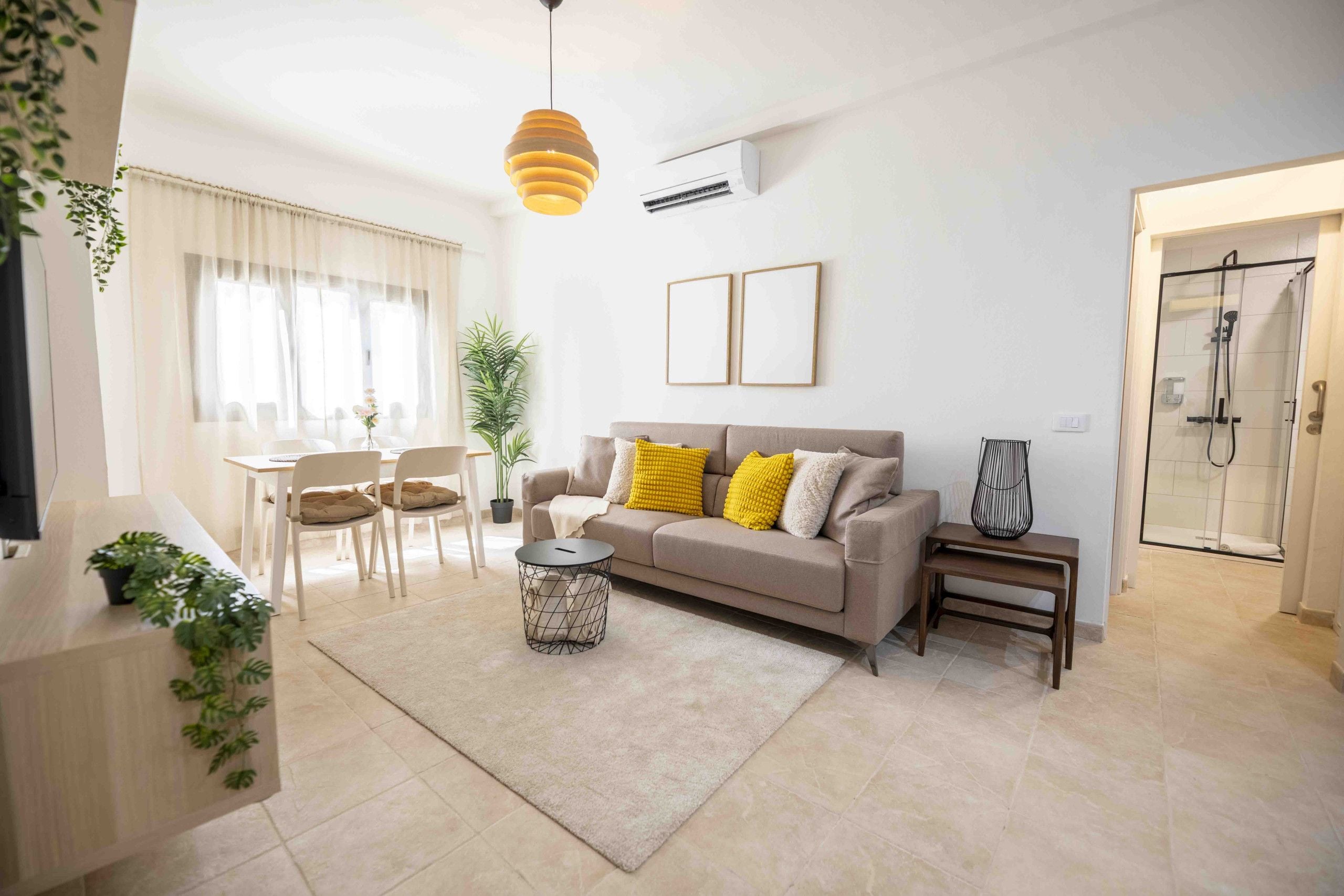 Una sala moderna con sofá, mesa de centro y sillas. Se observa un comedor con mesa y sillas. En el fondo, se ve una zona de ducha.