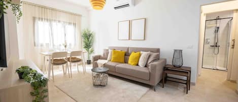 Una sala moderna con sofá, mesa de centro y sillas. Se observa un comedor con mesa y sillas. En el fondo, se ve una zona de ducha.