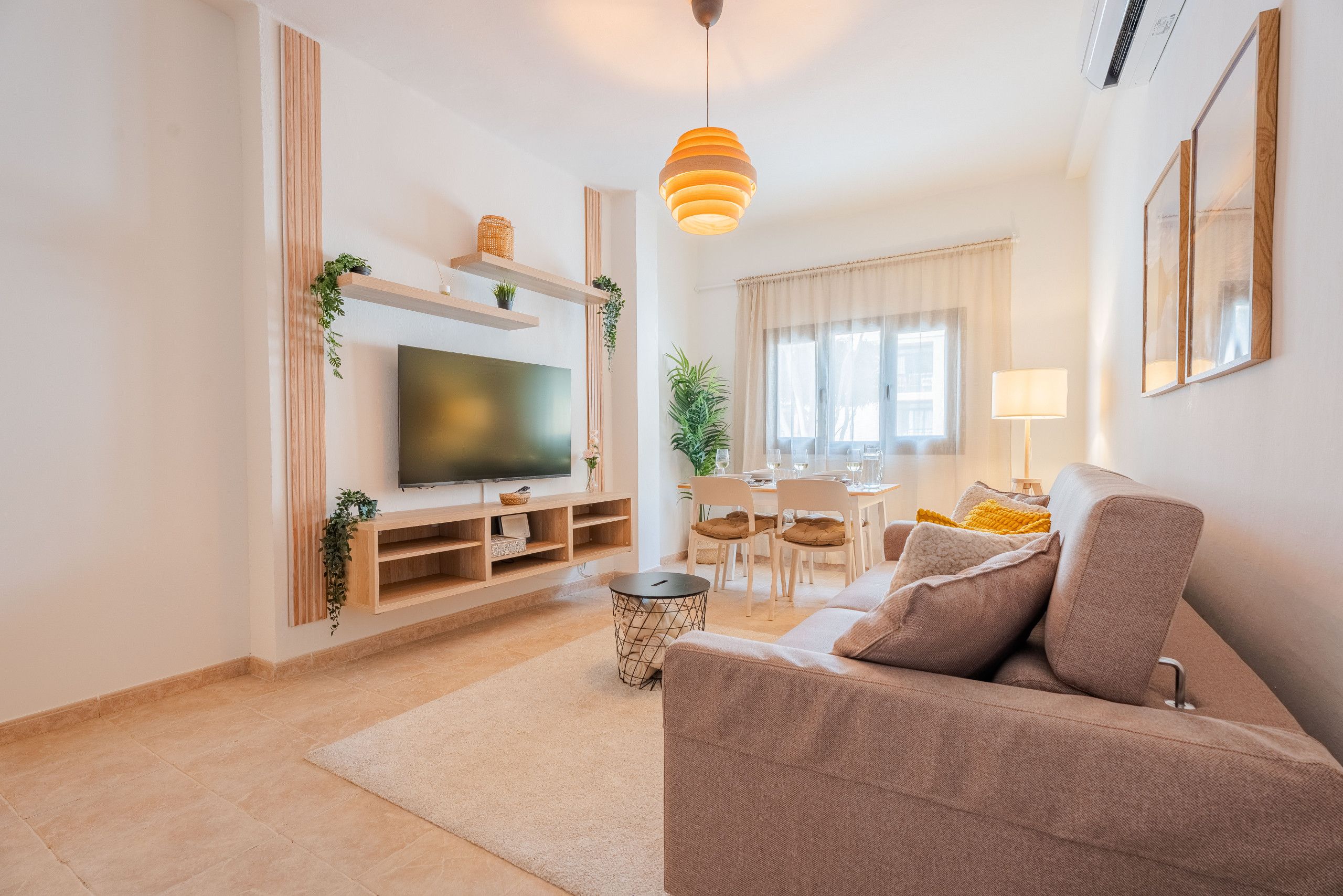 Een moderne woonkamer met een flatscreen televisie, houten planken, een eethoek met een tafel en stoelen, en een bank met kussens.