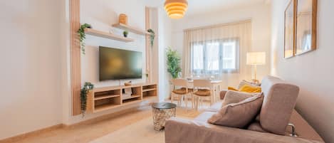 A modern living room with a flat-screen TV, wooden shelving, a dining area with a table and chairs, and a sofa with cushions.