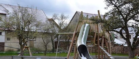 Un parque infantil con tobogán y estructuras para escalar, rodeado por una valla y árboles.