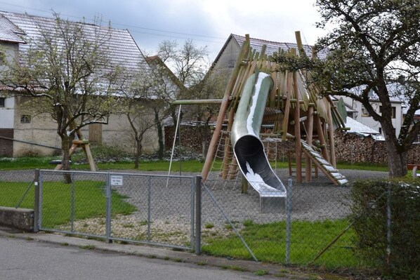 A playground with a slide and climbing structures, surrounded by a fence and trees. - Holiday home in Hülben with swimming pool (Hülben)
