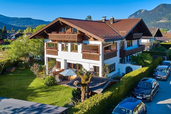 A two-story chalet with a sloped roof, wooden balconies, and a garden with a tree and a bench. - Alpklang Apartment #2 (Garmisch-Partenkirchen)