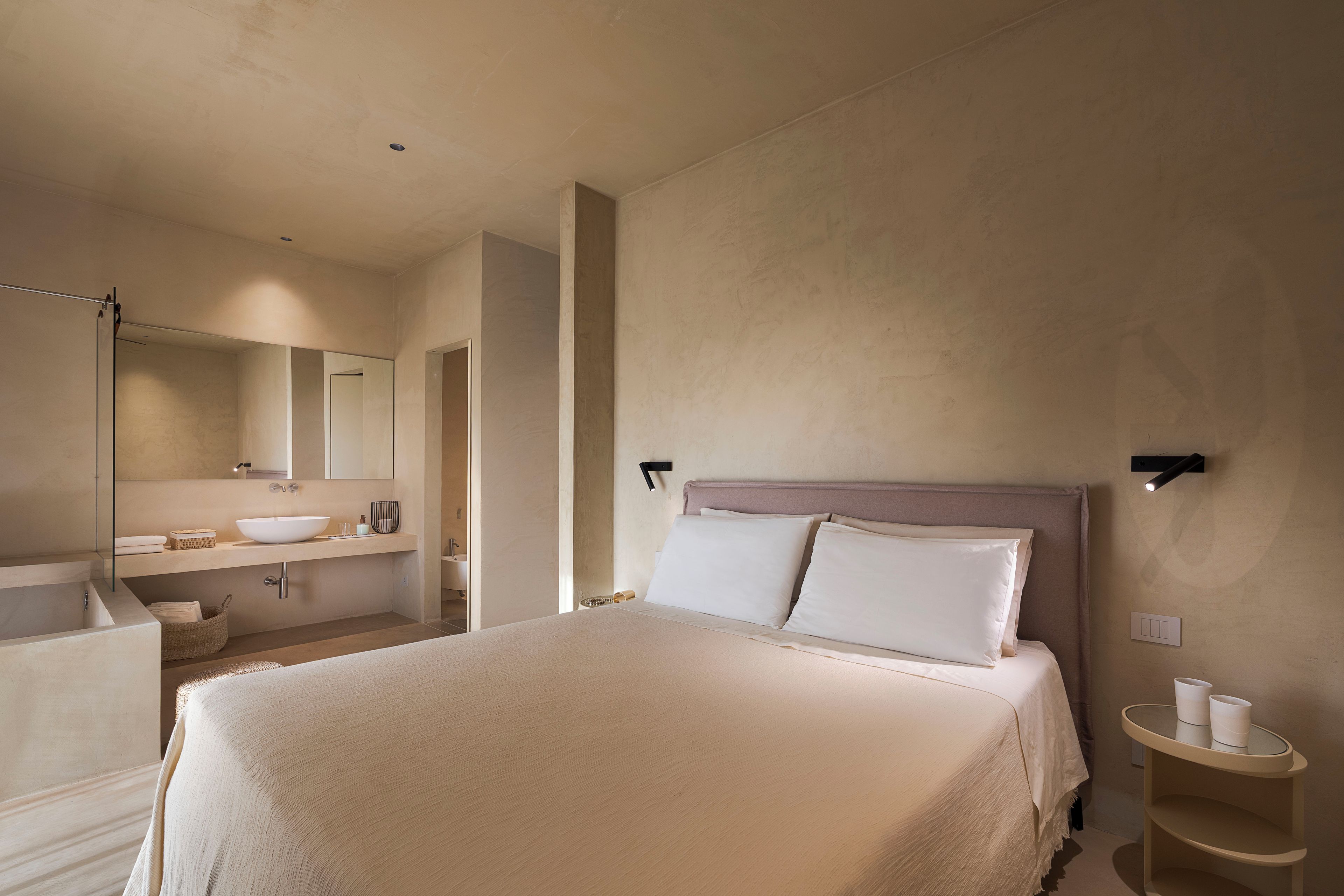 A modern hotel room with a large bed, a bathroom with a sink and mirror, and a small bedside table with cups.